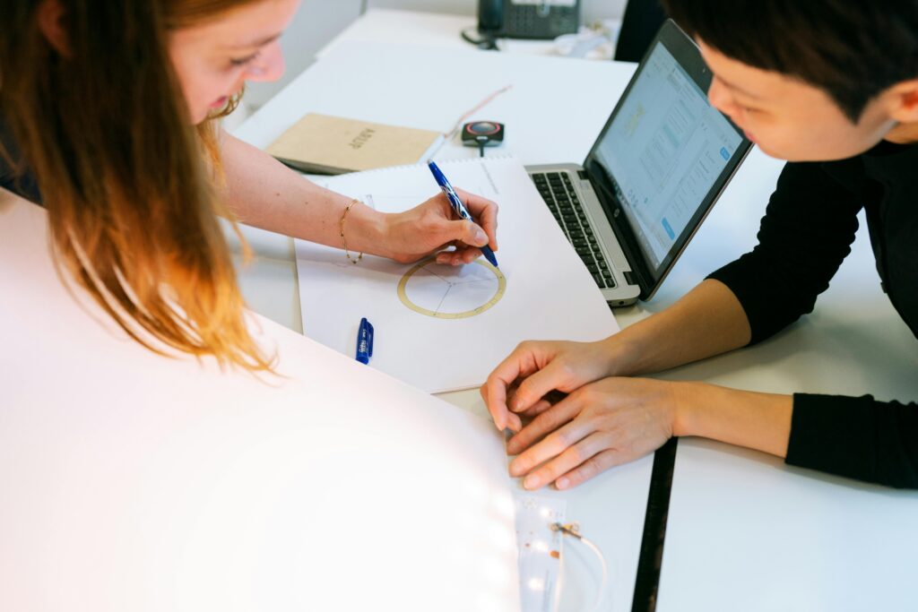 Two women collaborate on a design project at their desk, focusing on drawings and laptop.